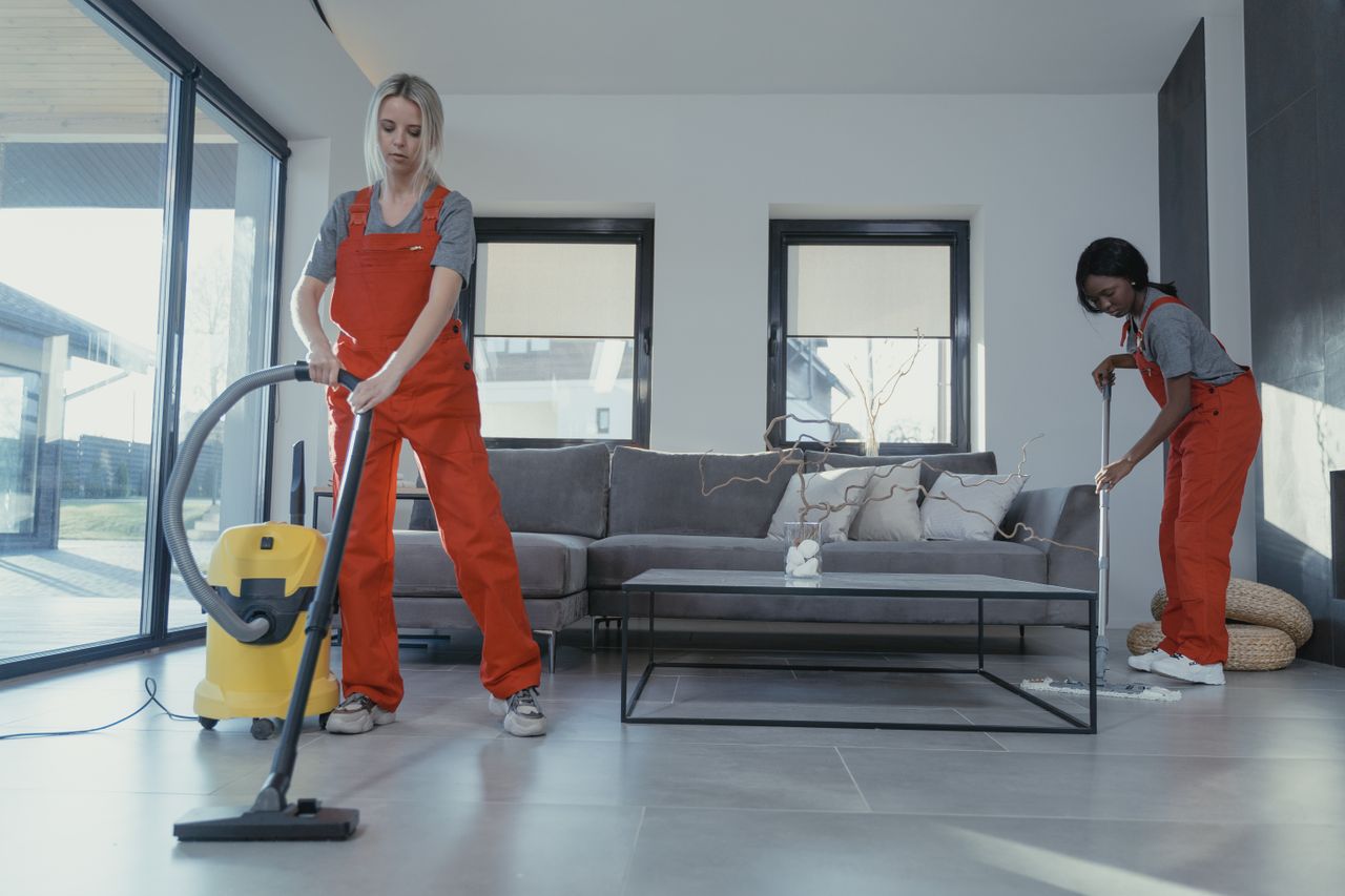 who-we-are Two women in red overalls vacuuming and sweeping a modern living room.