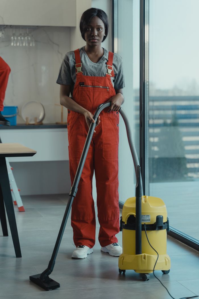services-03 An adult woman in red coveralls operates a vacuum cleaner in a modern indoor setting, focusing on cleaning tasks.